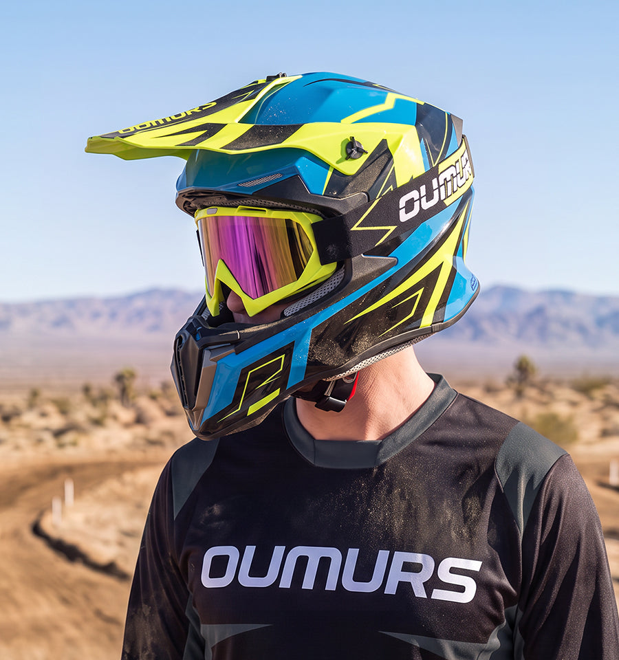 A person outdoors wearing a blue, yellow, and black full-face motocross helmet with a reflective purple-tinted visor, paired with a black OUMURS jersey. The background shows a blurred desert landscape under a clear blue sky.