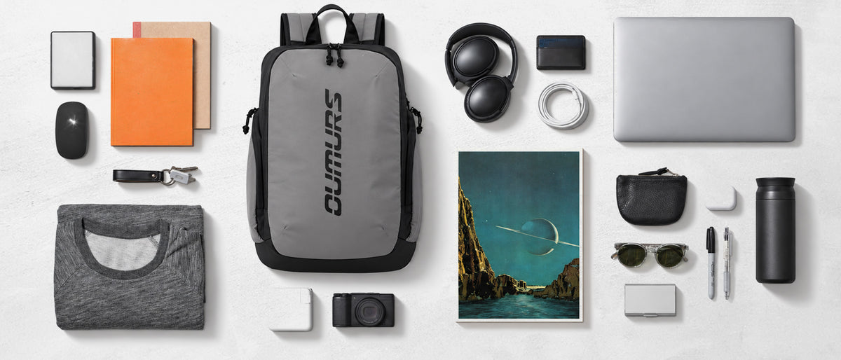 A gray and black DUMURS backpack is centered in a styled flat-lay arrangement with various tech accessories, notebooks, sunglasses, and a t-shirt on a light gray background.