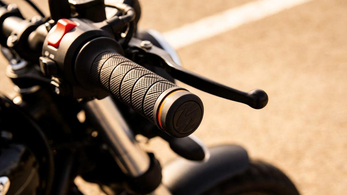Close-up view of a black ergonomic motorcycle handlebar grip featuring diamond pattern texture, orange accent band, and brake lever outdoors