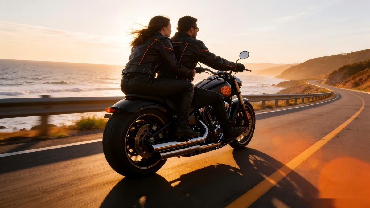Rear view of a couple in black leather jackets with orange trim riding a cruiser motorcycle on a coastal highway at sunset.
