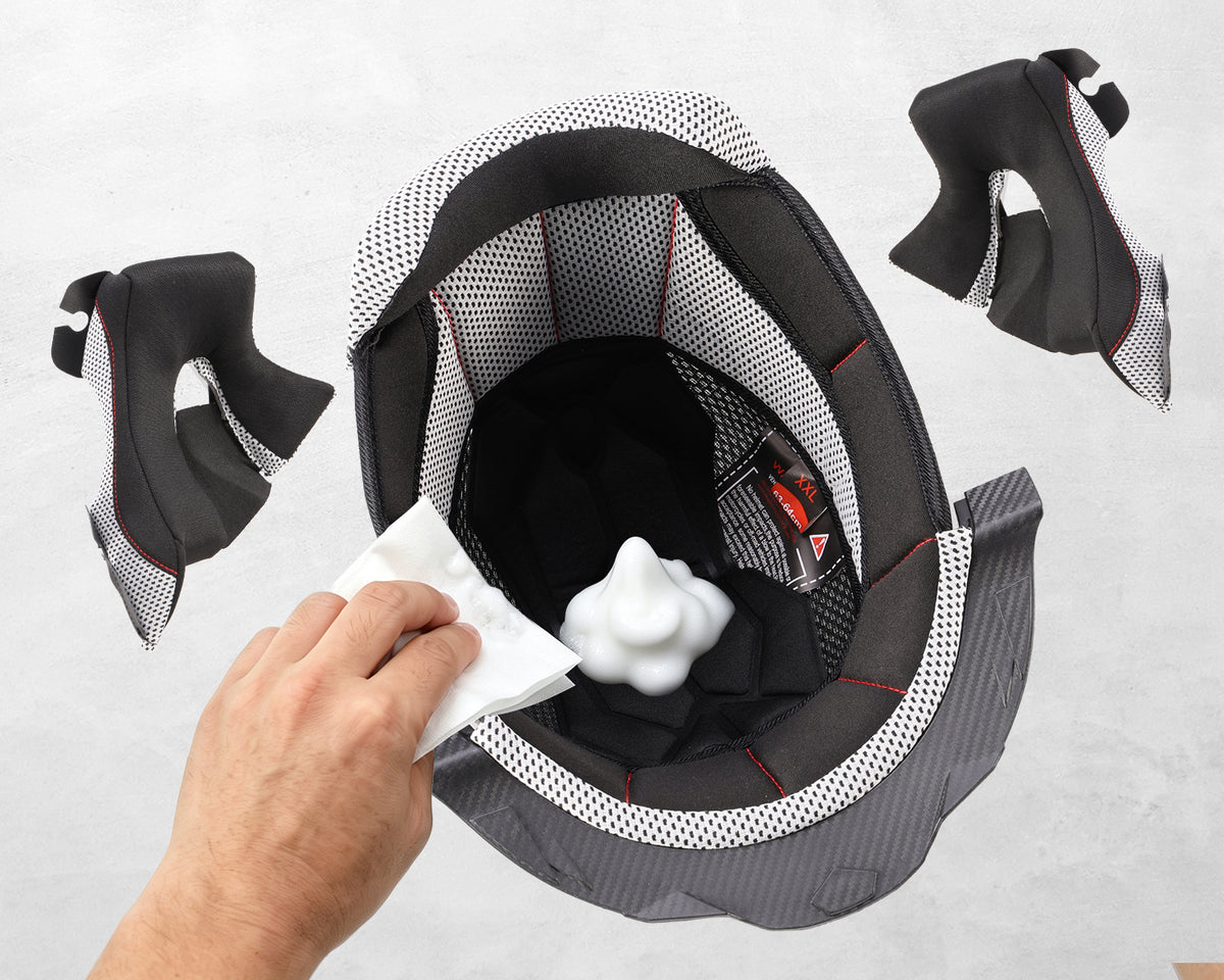 A helmet interior cleaning demonstration with a hand wiping the inside of a black and gray helmet with a white cloth, showing a white foam insert and black mesh lining.