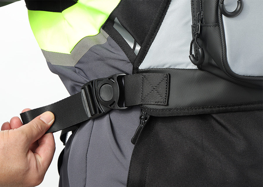 Close-up of a person fastening a black adjustable waist or hip strap with a plastic quick-release buckle onto a gray and black technical garment. A hand is visible on the left, and part of a neon yellow-green reflective section is visible at the top.