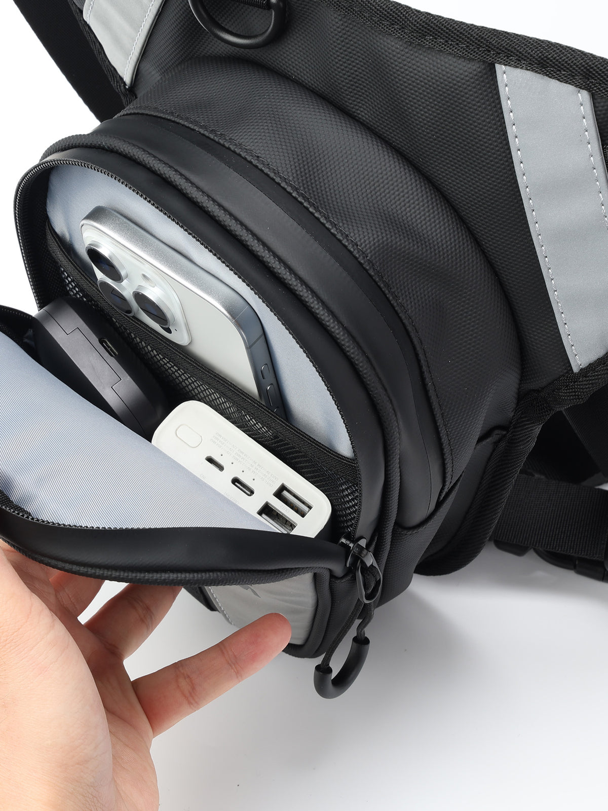 A close-up of a black and gray motorcycle leg bag with a hand opening the front zip pocket to show a phone and power bank inside. The bag has reflective gray side panels and a black nylon texture.