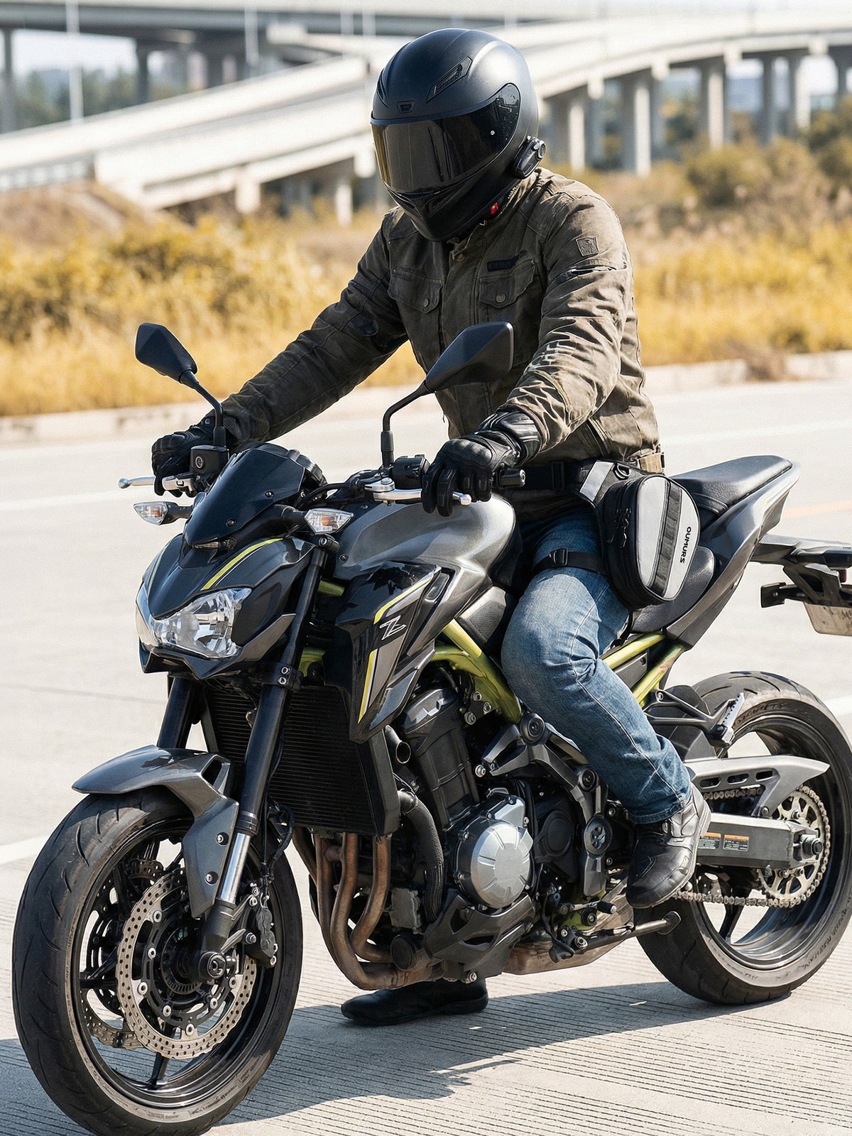 A rider wearing a black full-face helmet, olive green jacket, and blue jeans sits on a black and yellow Kawasaki ZX21R sport motorcycle on an outdoor paved road with a highway bridge in the background. wearing oumurs reflekta leg bag