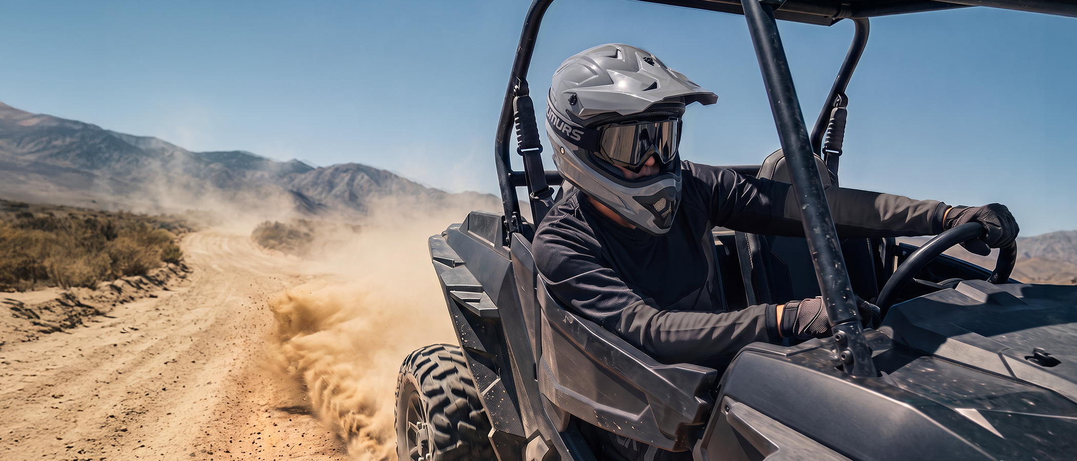 A person wearing an Oumurs 805 gray off-road helmet drives a black UTV on a desert road with mountains in the background.