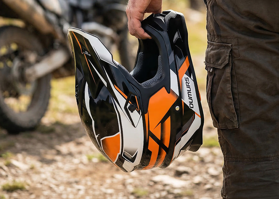 A close-up of a person holding a full-face motocross helmet. The helmet is black, white, and orange with the Dumura brand name visible on the side.