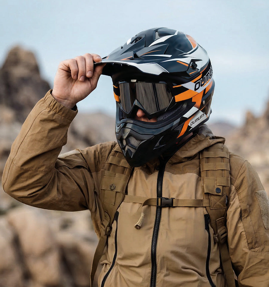 A person outdoors wearing a black, white, and orange full-face motorcycle helmet with tinted goggles, a tan jacket, and a backpack, with blurred rocky terrain in the background.