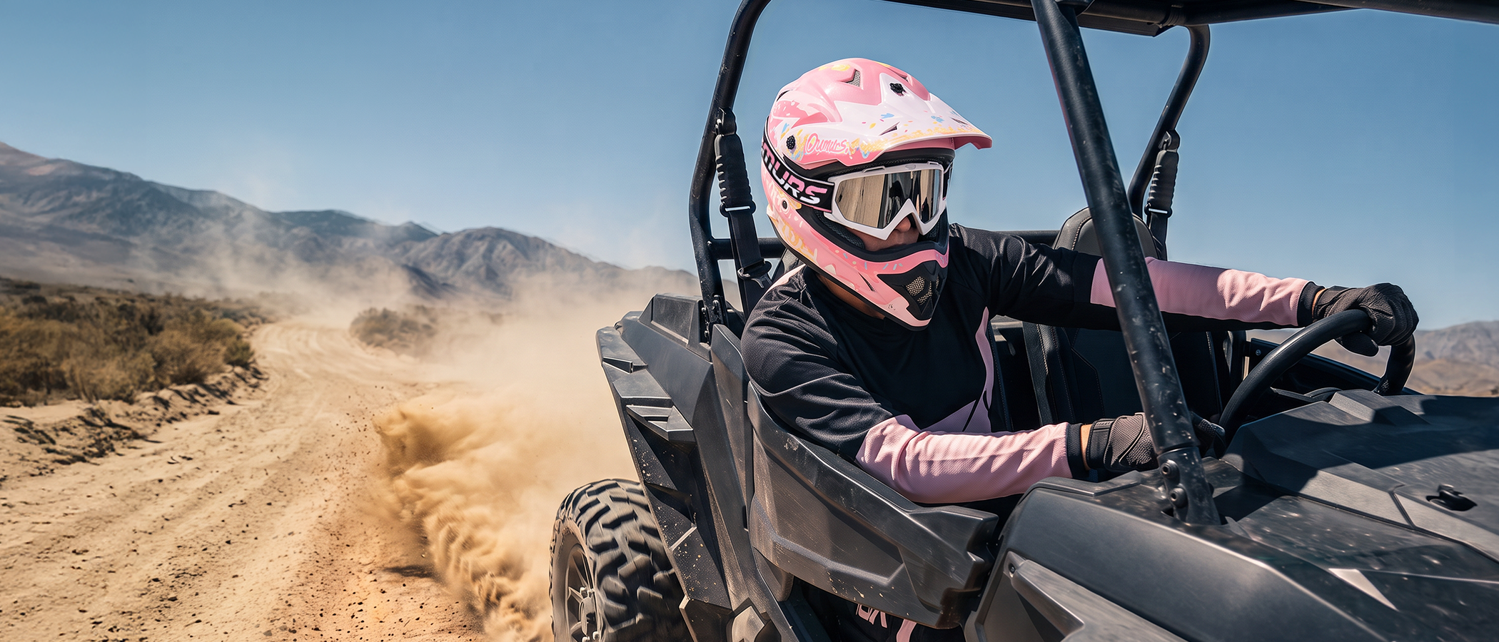 A person wearing an Oumurs 805 pink off-road helmet drives a black UTV on a dusty dirt trail with mountains in the background.