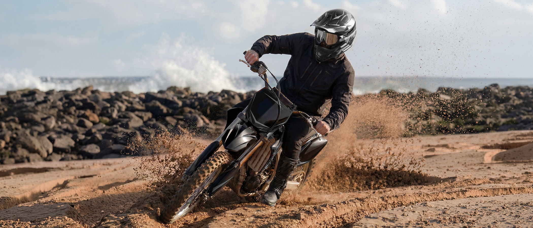 A rider wearing an Oumurs 805 Victory Gray off-road helmet leans into a turn on a dirt bike along a rocky coastline, with waves crashing in the background.