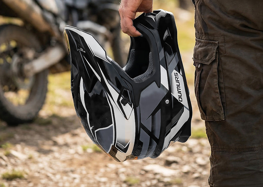 A rider holds the Oumurs 805 victory gray off-road helmet in one hand while walking on a rocky trail.