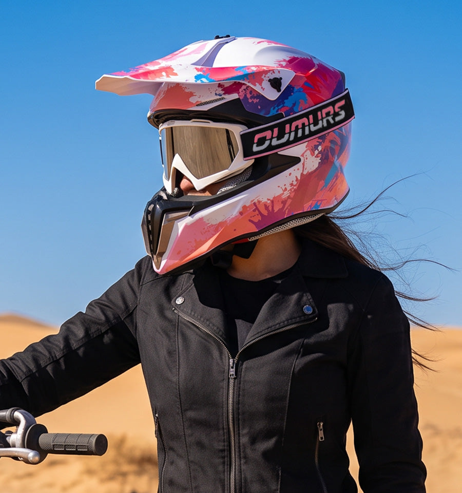 A person wearing a pink, white, and blue graphic full-face motocross helmet with a visor and goggles, riding a motorcycle in a desert landscape under a clear blue sky.
