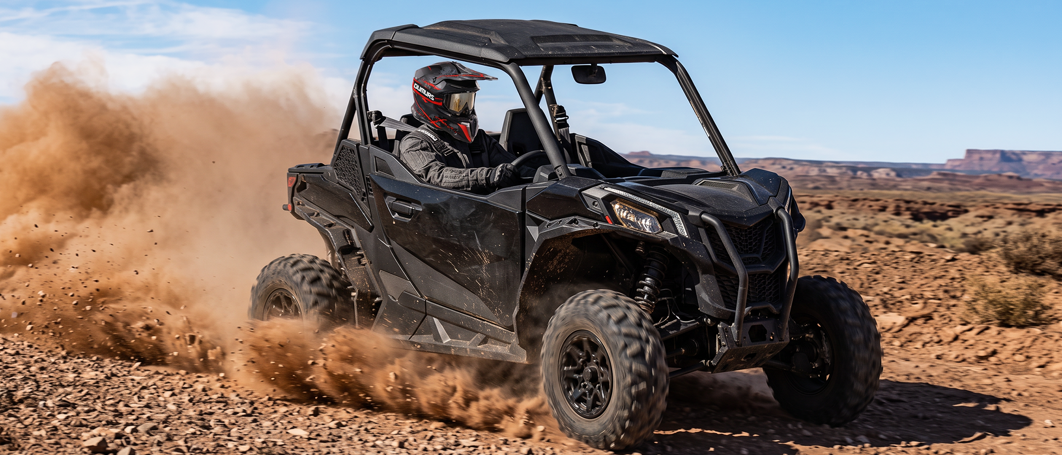 A person wearing an Oumurs 806 Red Skull off-road helmet drives a black UTV on a dusty dirt trail while kicking up a large cloud of dust. 