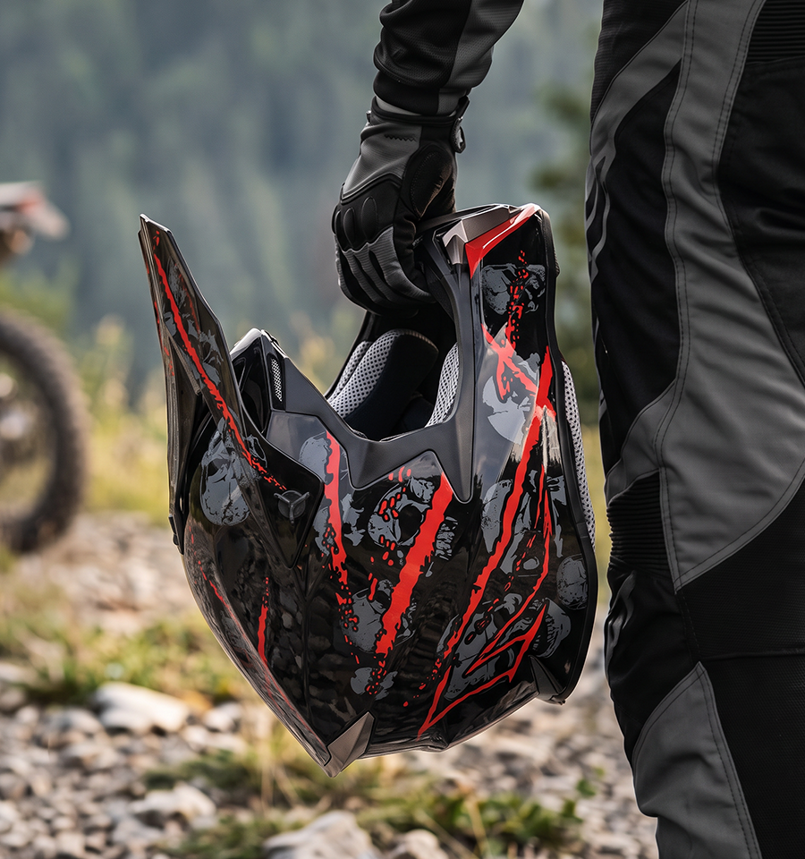 A rider holds the Oumurs 806 Red Skull off-road helmet in one hand while walking on a rocky trail.