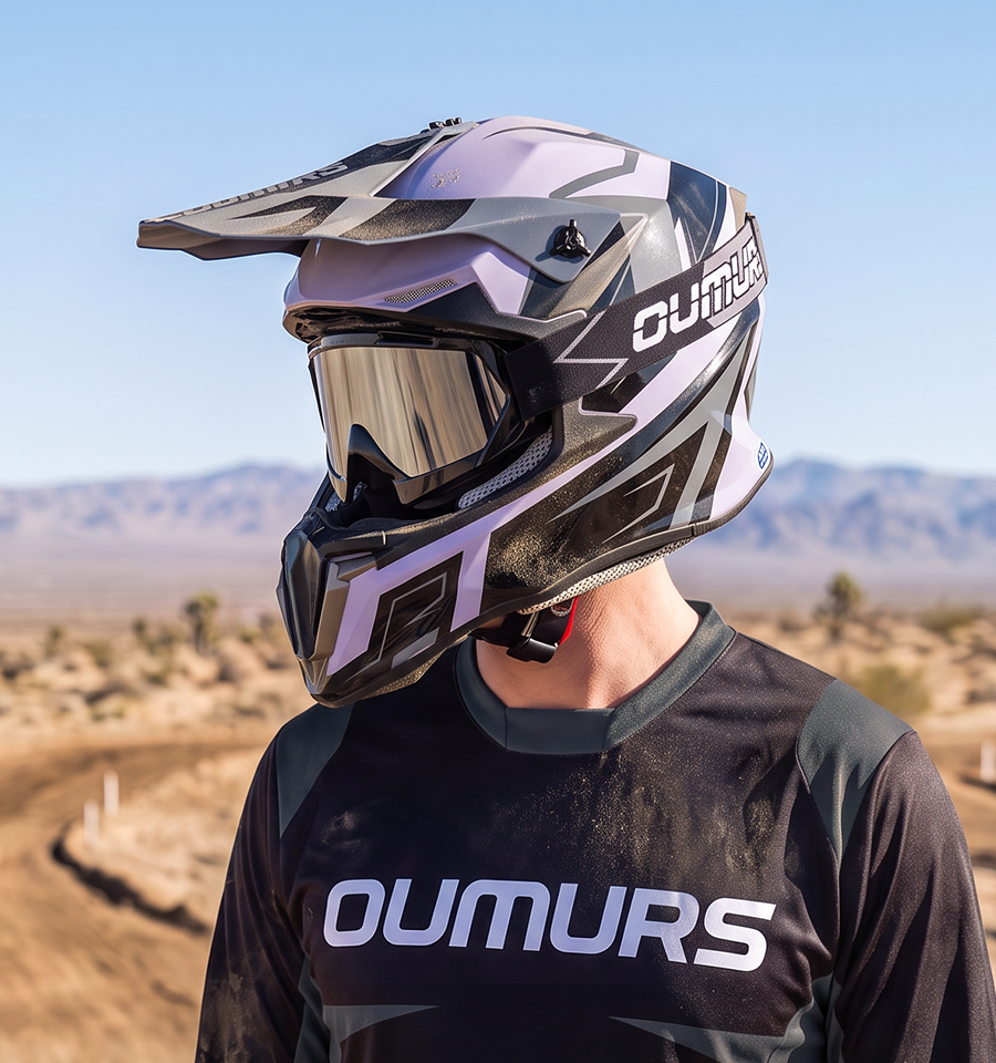 A person outdoors in a desert landscape wearing a full-face off-road helmet and a matching black and gray OUMURS jersey. The helmet has a black, white, and light purple/lavender color scheme with a dark mirrored visor.