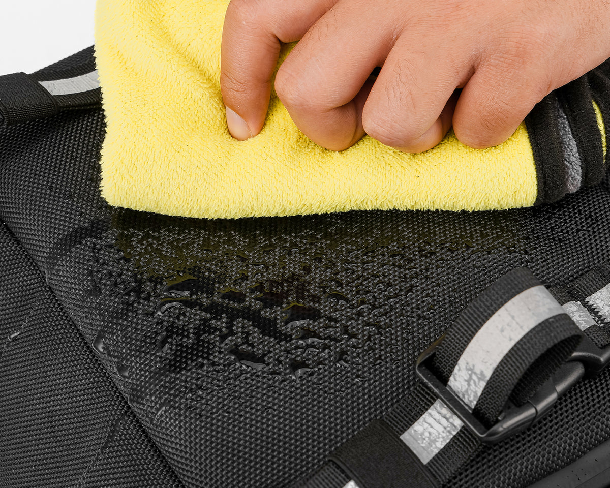 Close-up of water droplets on black backpack fabric being wiped with a yellow cloth.