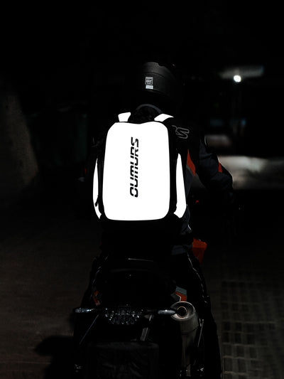 A person wearing a black motorcycle helmet and riding gear sits on a motorcycle at night, viewed from behind. The rider is wearing a highly visible white backpack with reflective white side panels and the brand name 'CUMURS' printed vertically in black on the back.
