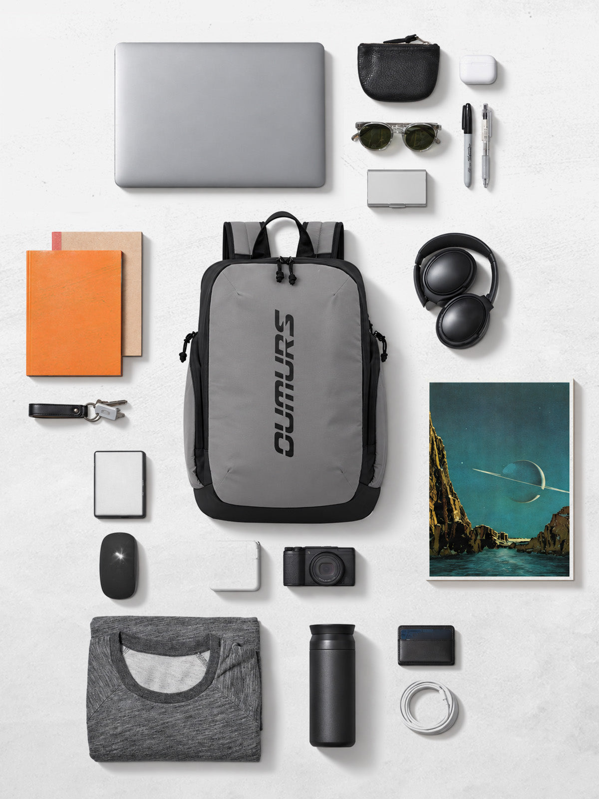 A gray and black Clumsy backpack is centered in a styled flat-lay arrangement on a light gray surface, surrounded by various tech and lifestyle accessories including a laptop, headphones, camera, notebook, and framed art print.