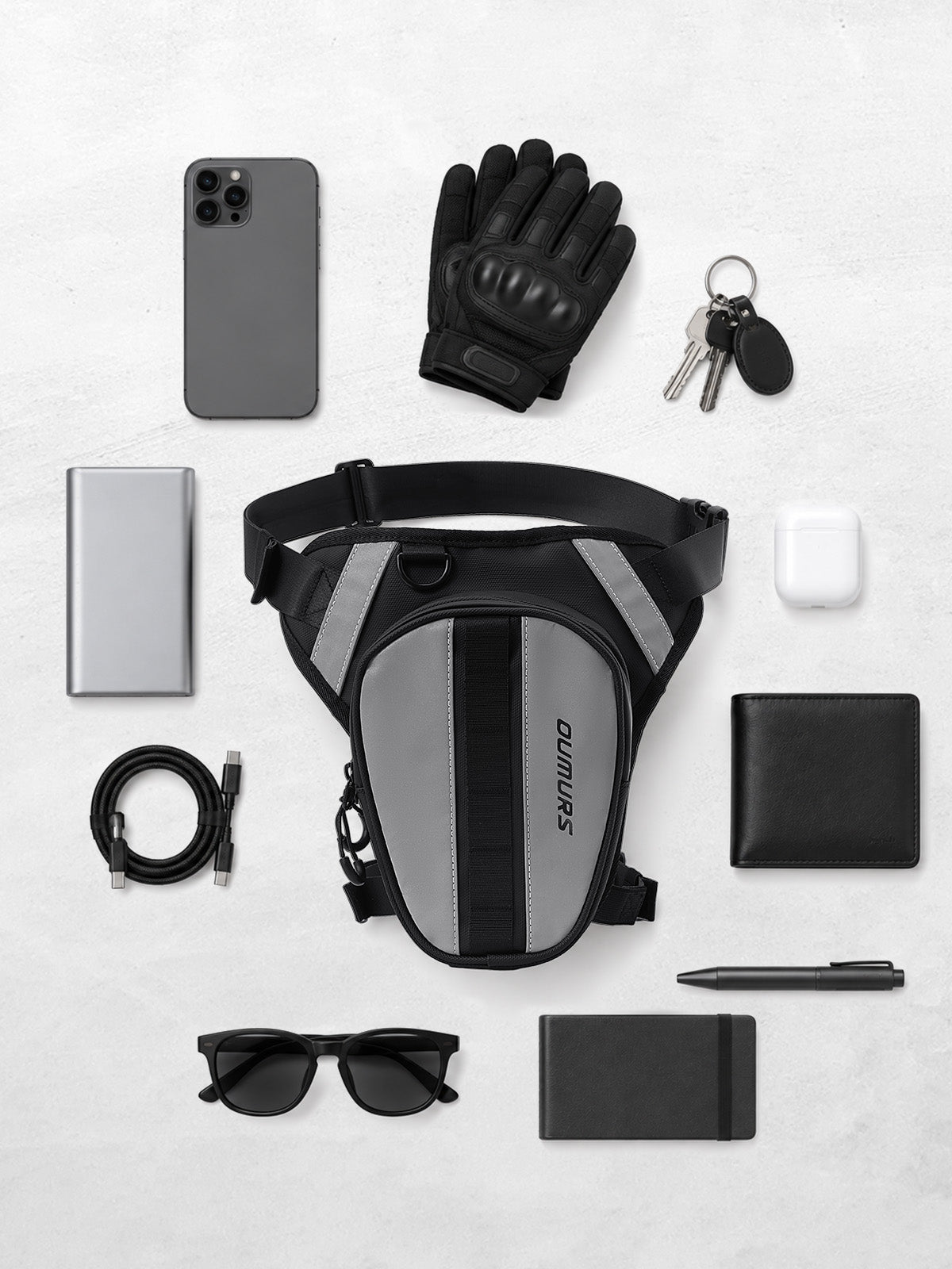 Black and gray Oumurs leg bag surrounded by a phone, gloves, keys, wallet, sunglasses and other small essentials.