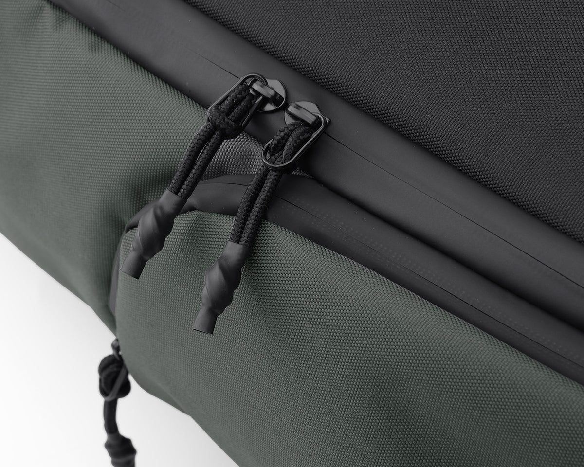  Close-up of the Oumurs backpack’s dual zipper pulls and covered zipper seam on dark green fabric.