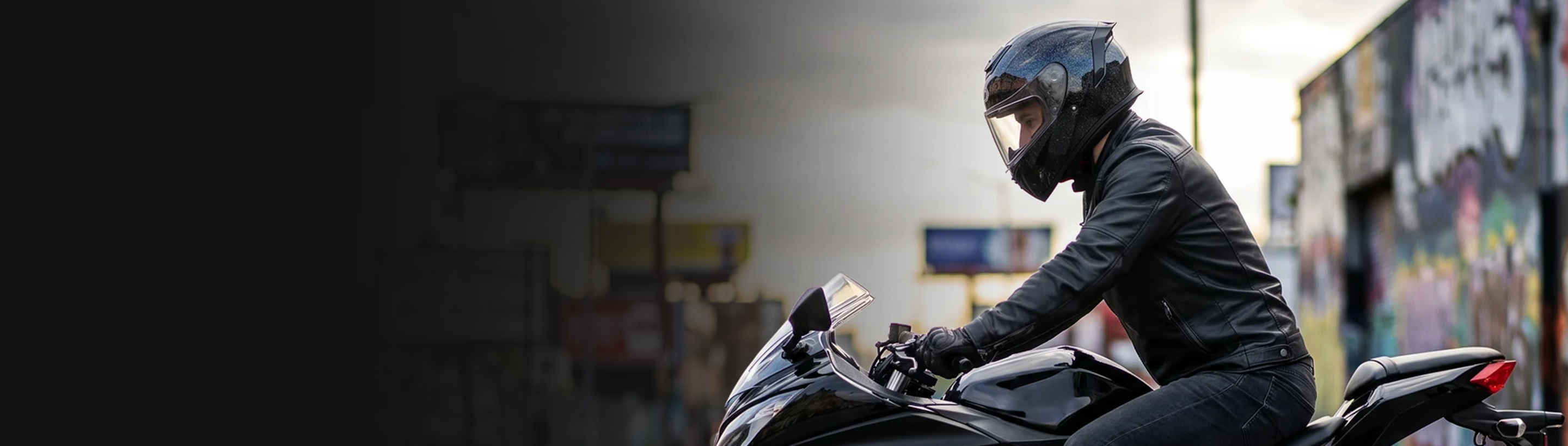 A person wearing a cosmo black Oumurs 361S helmet rides a black motorcycle in a city street with blurred buildings and graffiti in the background.