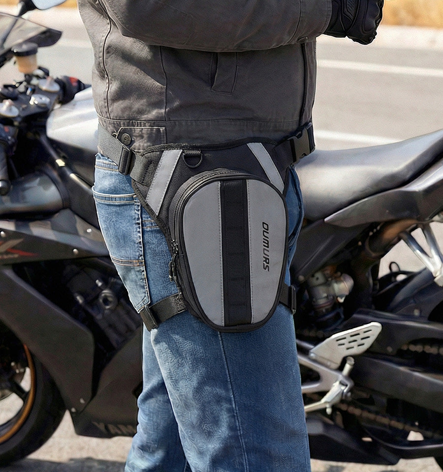 Rider wearing a black and gray Oumurs leg bag secured around the waist and thigh beside a motorcycle.