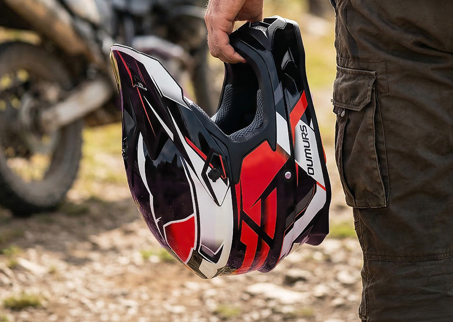 A person holds a black, red, and white full-face off-road motorcycle helmet with Dumita branding, set against a blurred outdoor background with dirt, gravel, and a motorcycle.