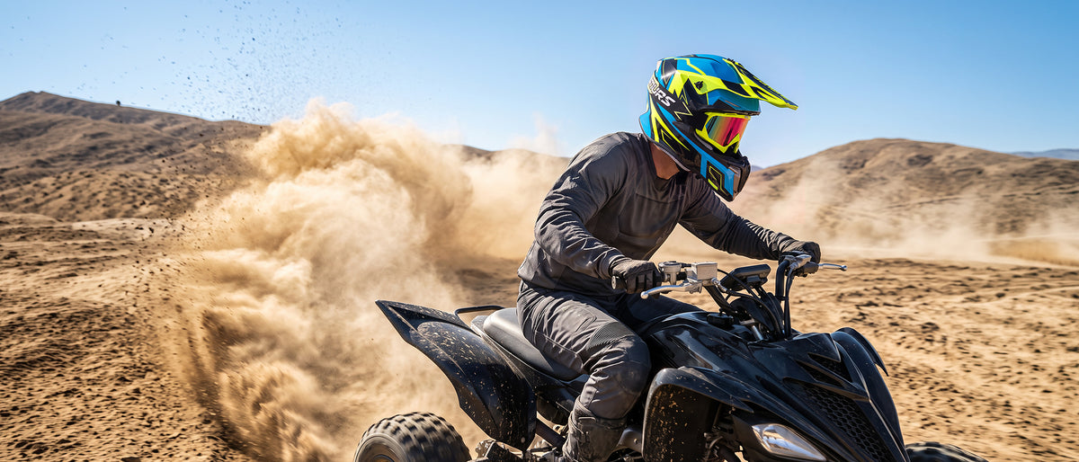 A person wearing a blue, yellow, and black full-face helmet and dark riding gear operates a black quad bike in a dusty desert landscape, kicking up a large cloud of sand under a clear blue sky.