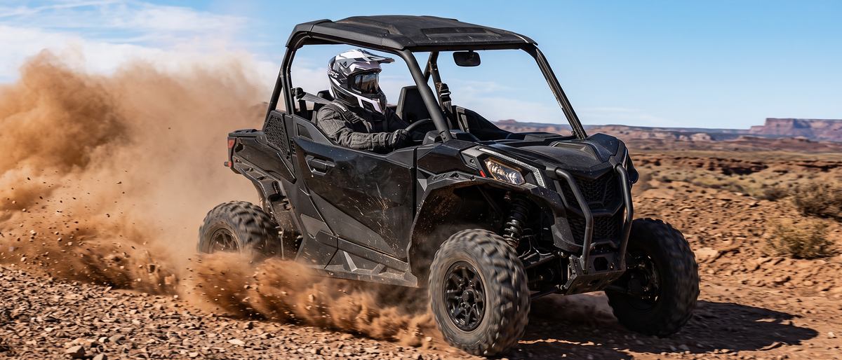 A black off-road side-by-side UTV with a roll cage and canopy, driven by a person in a desert landscape and kicking up a large cloud of dust.